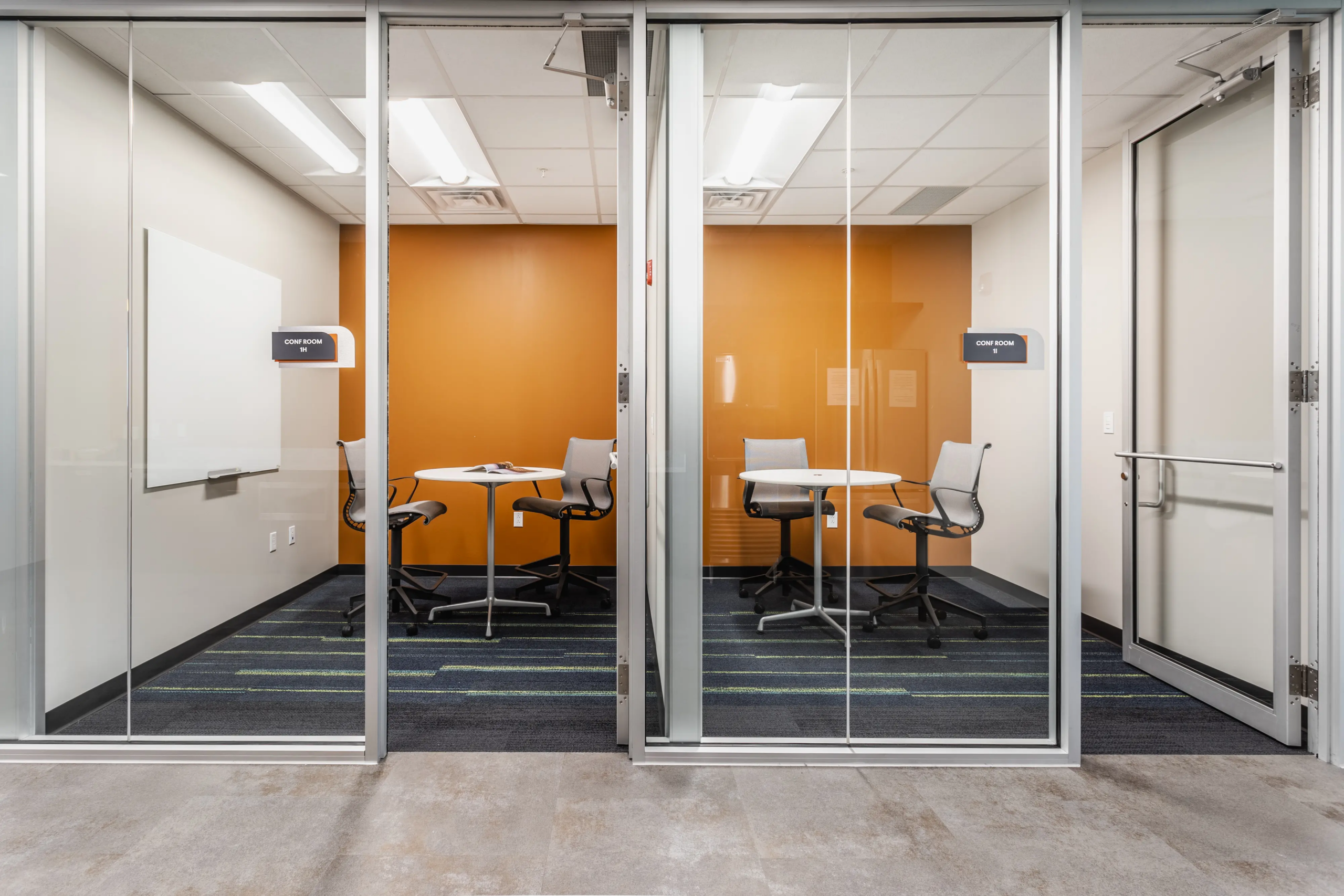 Glass front huddle rooms with orange accent wall and small meeting tables in modern office